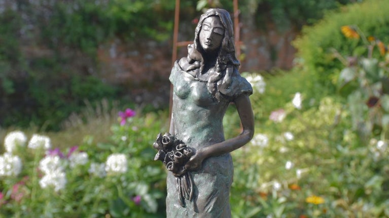 A sculpture of a woman holding a bunch of roses, set against a backdrop of colourful flowers and stone walls